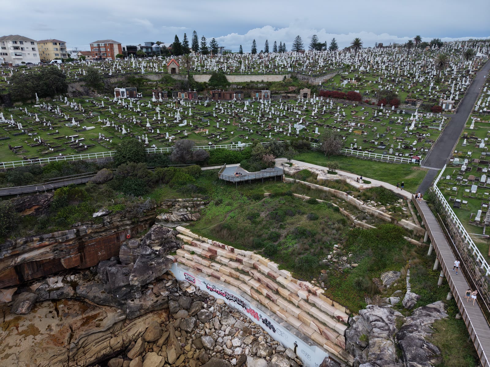 Aerial view of a clifftop cemetery above a rocky shoreline, a coastal boardwalk winding along the sea edge