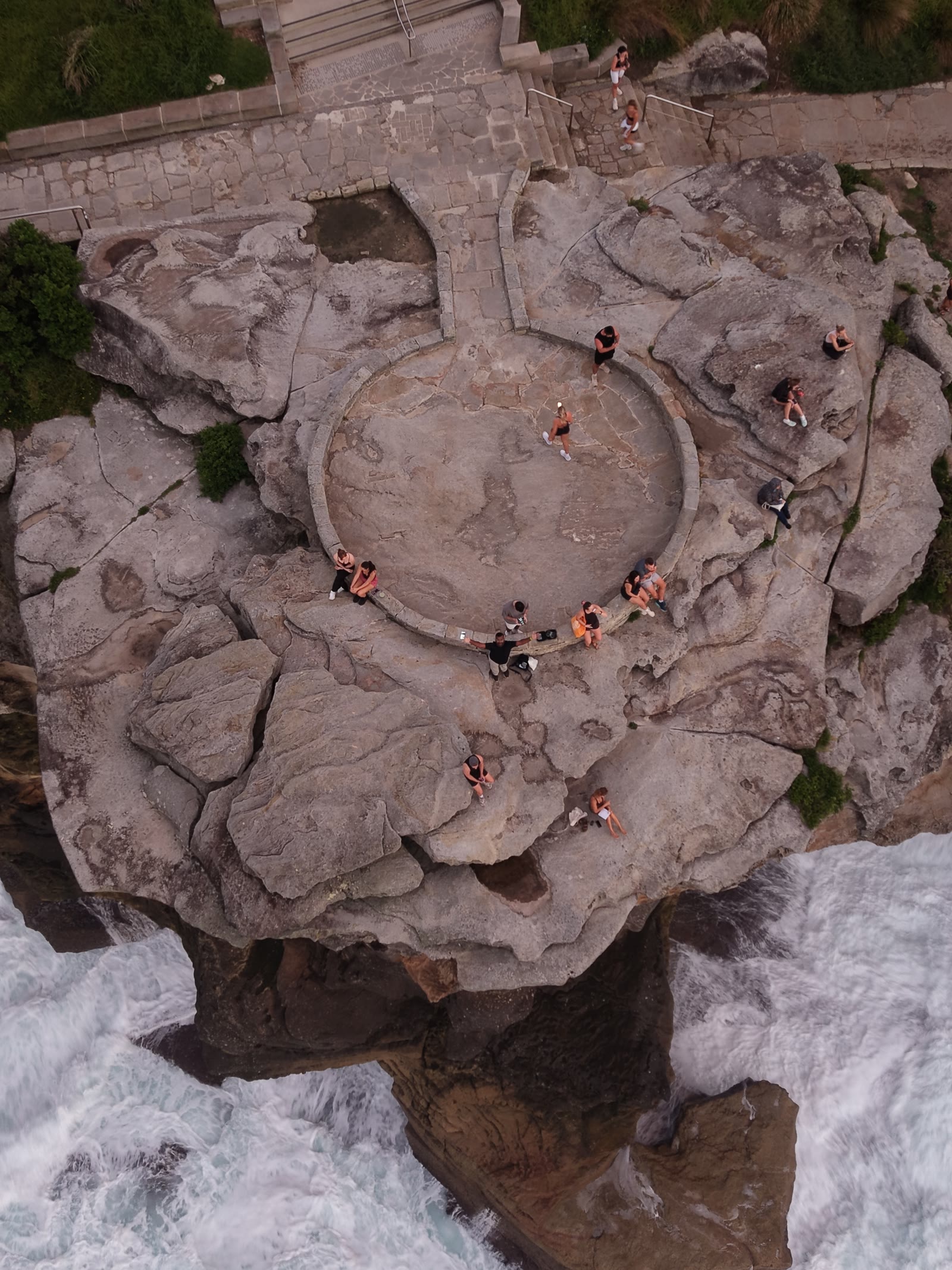 Top-down view of a circular clifftop lookout ringed with visitors, surf churning against the rocks beneath