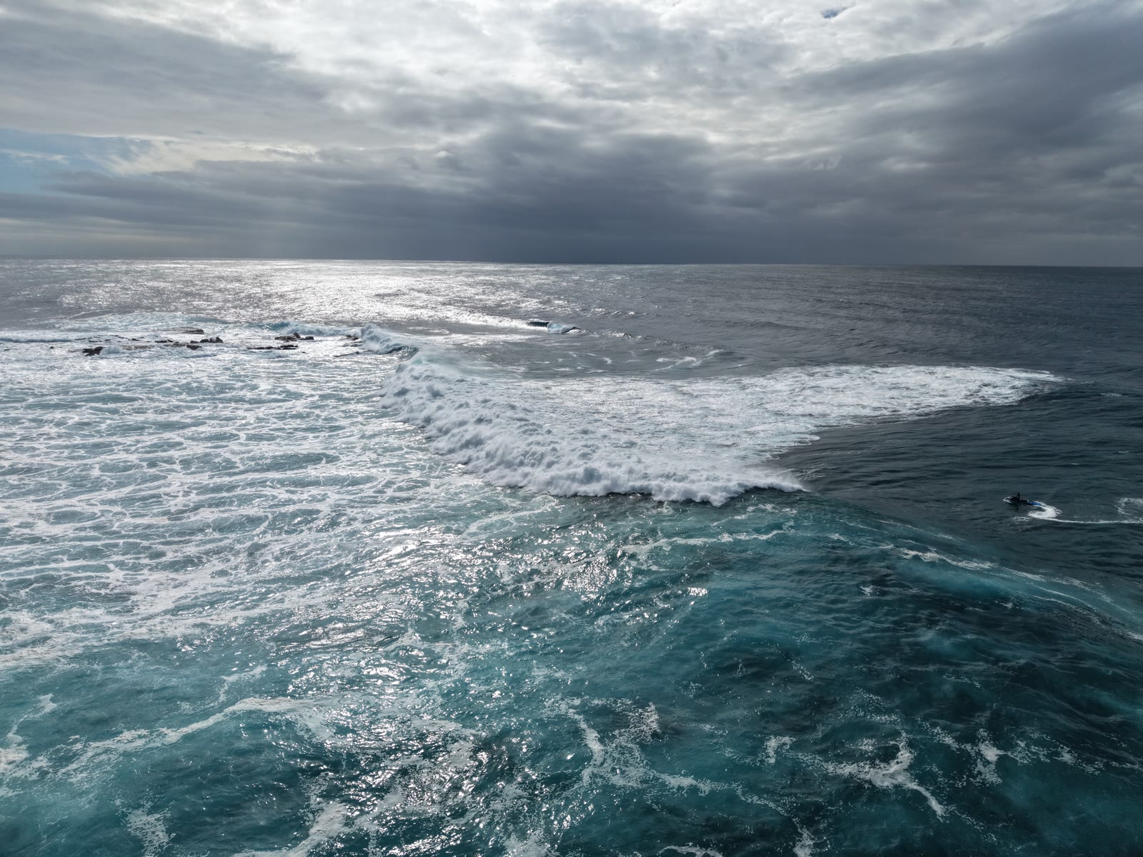 Storm light over the ocean as a wave breaks across a shallow reef, a lone jet ski waiting beyond the foam