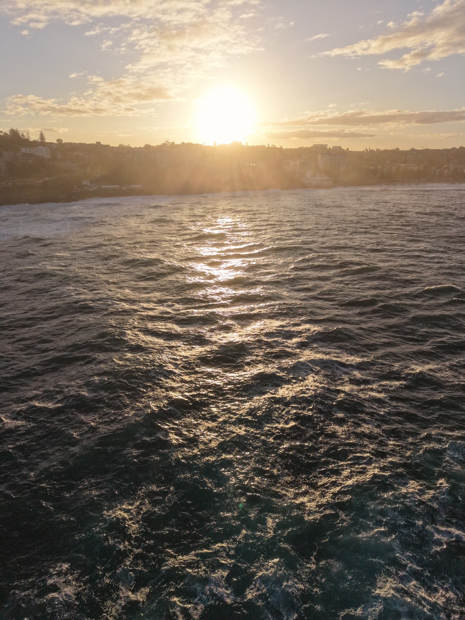Sunset over a silhouetted coastal suburb photographed from above the ocean, golden light on the swell