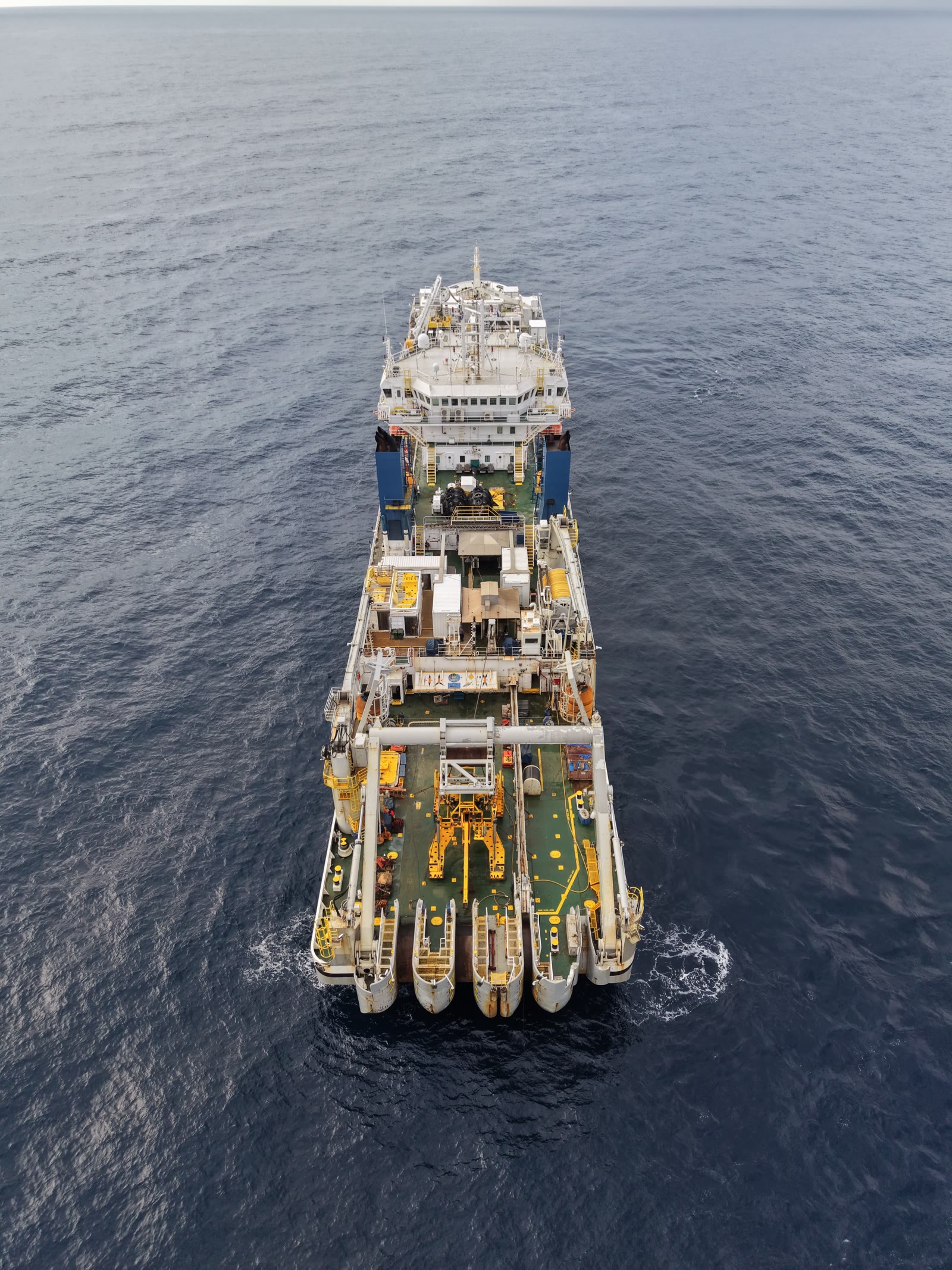 Straight-down aerial of an offshore work vessel mid-ocean, deck cranes and survey gear visible